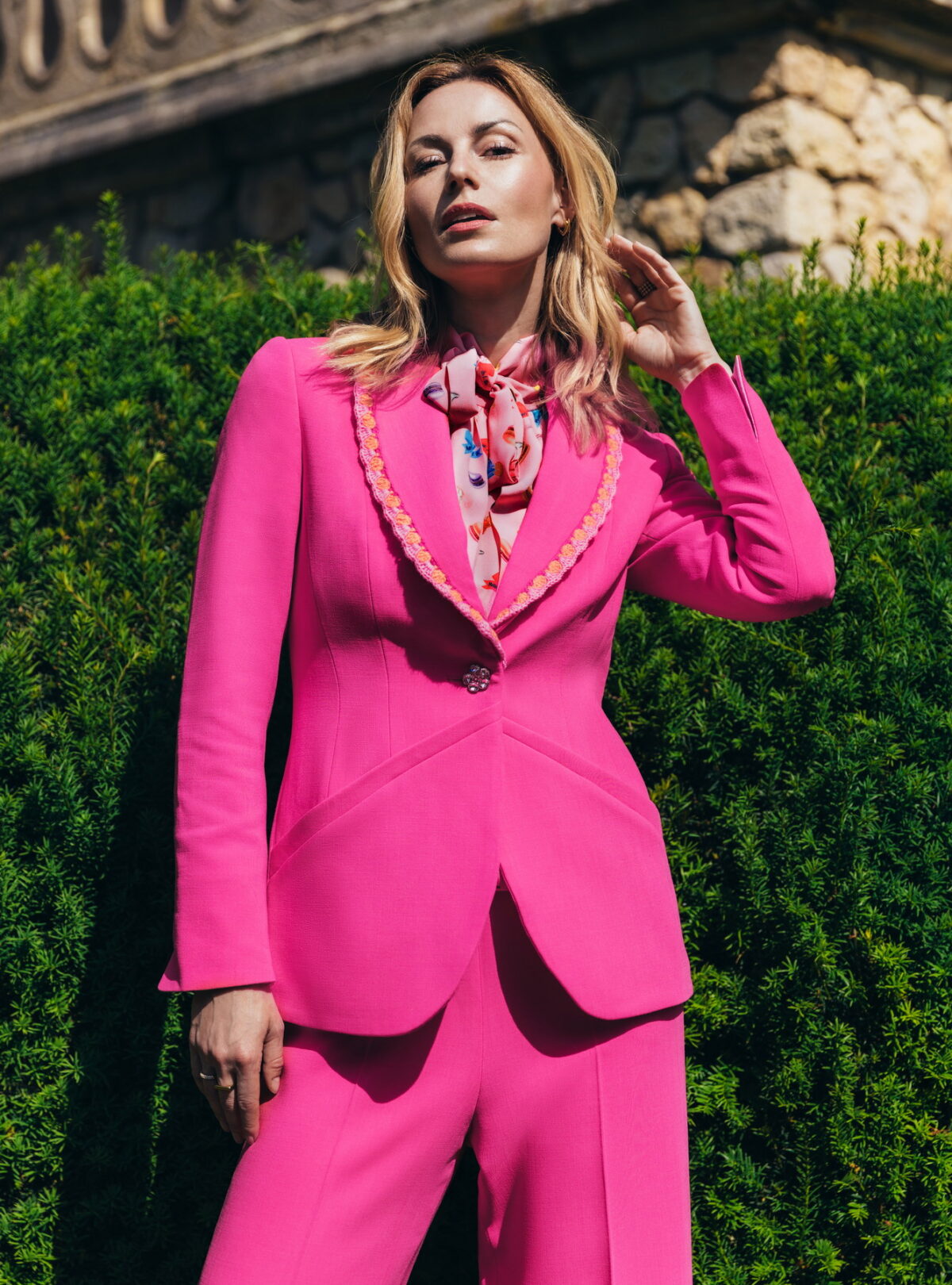 "Frau trägt pinken Blazer mit Häkelkante und Schalkragen von Maison de l’île – Close-up vor grüner Hecke"